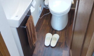 床のクッションフロアと背面の壁紙が木目のトイレ室内と手洗い器